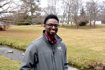 Austin Gray, wearing a Virginia Tech windbreaker over a sweatshirt, stands by Stroubles Creek on the Virginia Tech Blacksburg campus.  Behind him, in addition to the creek, are a long swath of grass, bare trees, and a house in the distance.